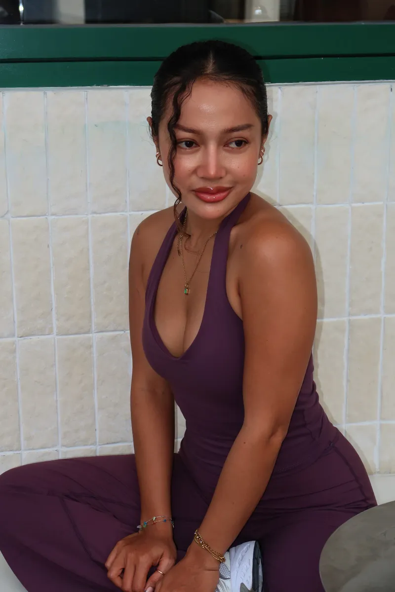 Jean in a plum halter top, sitting against a white tiled wall with a soft smile