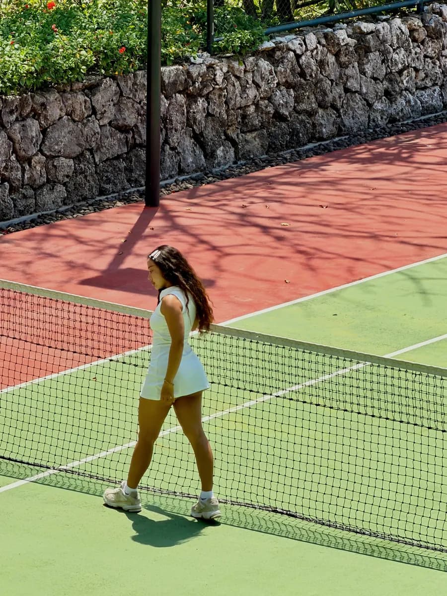 Jean in a white tennis dress walking across a tennis court in the sun