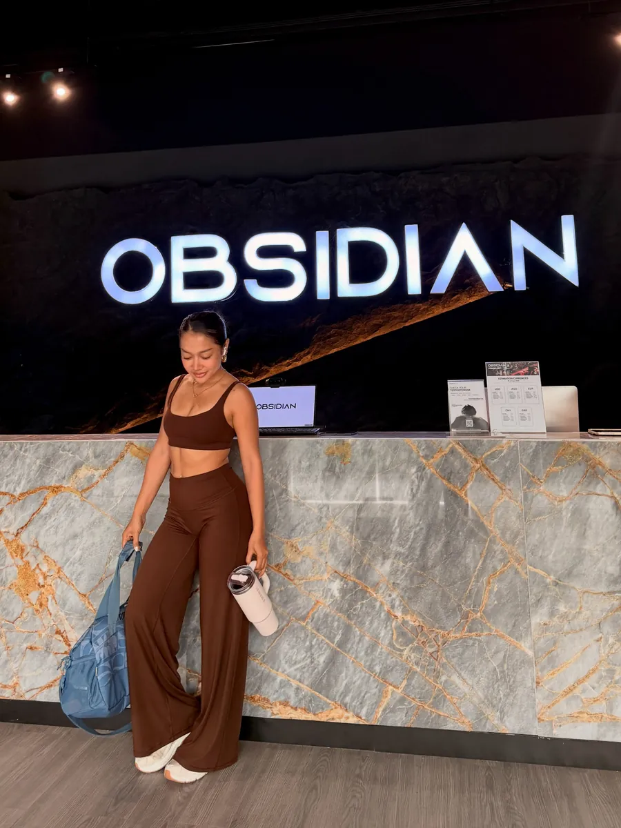 Jean in a brown training set with her gym bag, standing at the front desk of her gym